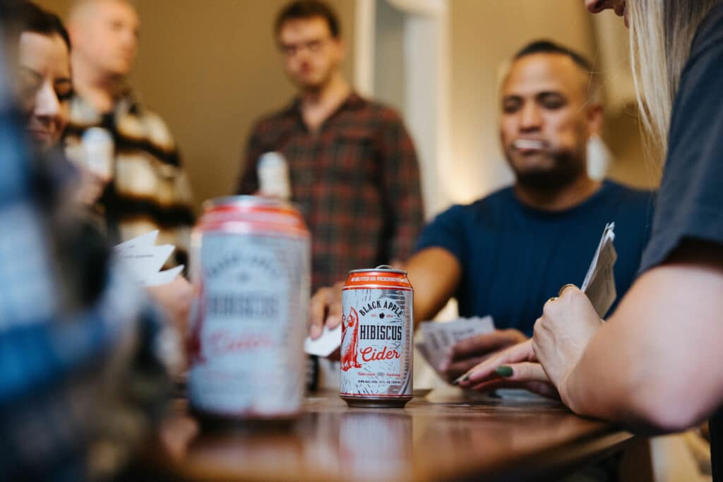 Photo of a group of friends playing cards, focused on a can of Black Apple Hibiscus hard cider.