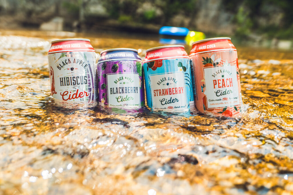 Cans of Black Apple Hibiscus, Blackberry, Strawberry, and Peach Cider all sitting in a creek.